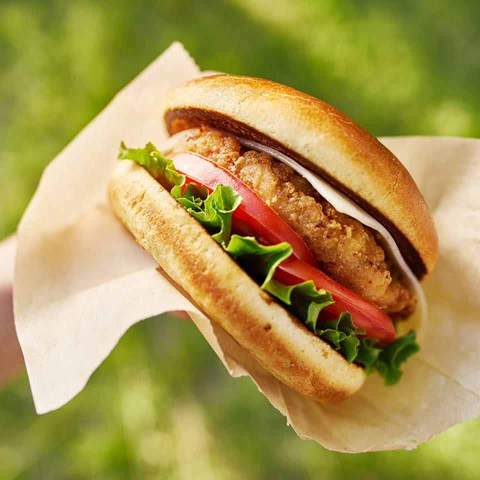 Crispy chicken sandwich with lettuce, tomato, and sauce served on parchment outdoors against a blurred green background.
